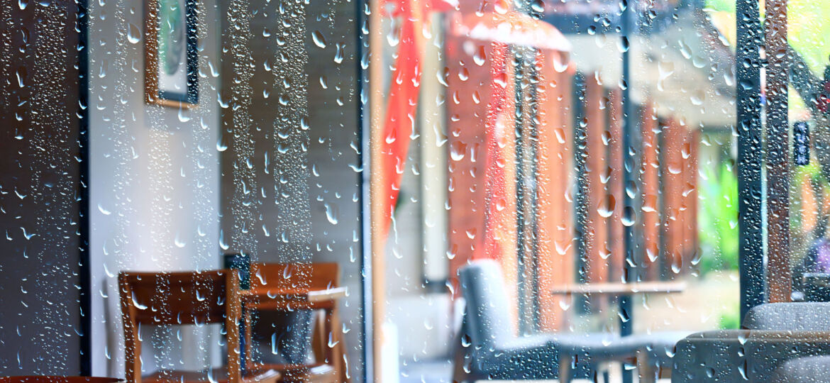 View through a raindrop-covered window overlooking a cozy café interior with empty wooden chairs and tables. The vibrant colors of the street and buildings outside are blurred, creating a soft bokeh effect. The raindrops add a tranquil, reflective quality to the scene, evoking a sense of calm during a rainy day.