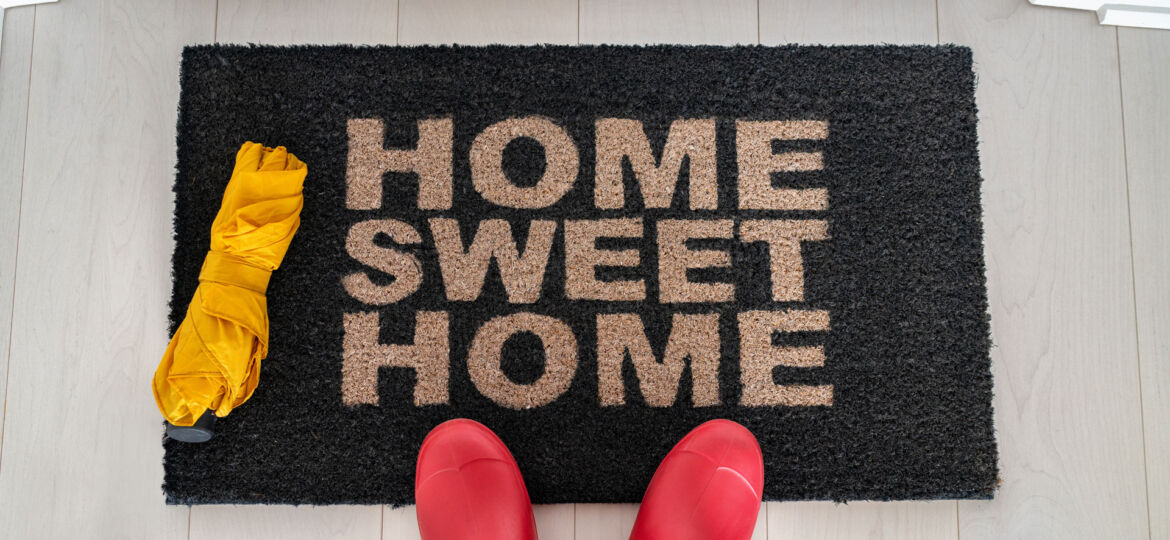 First-person perspective of a person standing on a black doormat with the welcoming phrase 'HOME SWEET HOME' written in beige letters. To the left of the mat is a folded yellow umbrella, and the person is wearing bright red rain boots, suggesting they have just arrived home on a rainy day.
