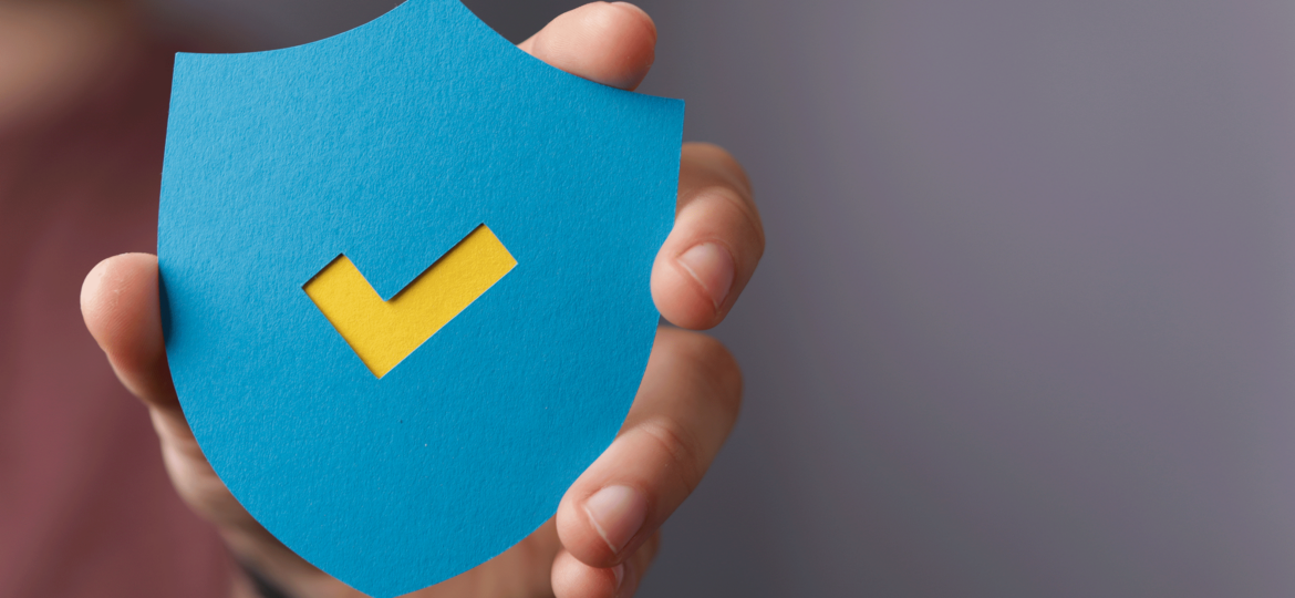 Hand holding a blue paper cutout in the shape of a shield with a yellow checkmark in the center. This is to represent the symbolic safety shield that epoxy coatings provide for concrete.