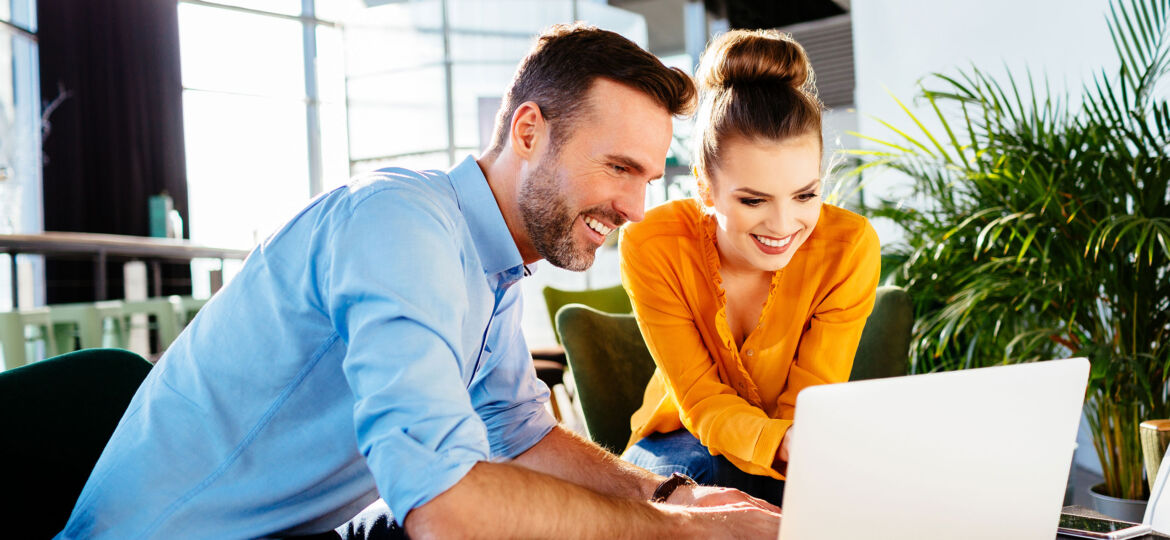 A man and woman sit together, smiling as they look at a laptop screen in a brightly lit modern office or cafe. The man, wearing a light blue shirt, types on the keyboard while the woman, in a vibrant yellow blouse with her hair tied in a bun, leans forward, appearing engaged in the discussion. Large windows and green plants are visible in the background, adding a natural and airy feel to the space.