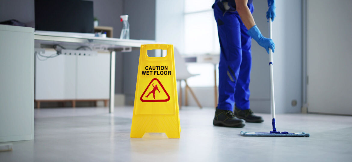 A janitor wearing blue gloves and a uniform mops a light-colored office floor. A bright yellow "Caution Wet Floor" sign with a warning icon is prominently placed in the foreground. The office background includes a desk, a chair, and cleaning supplies on a surface.