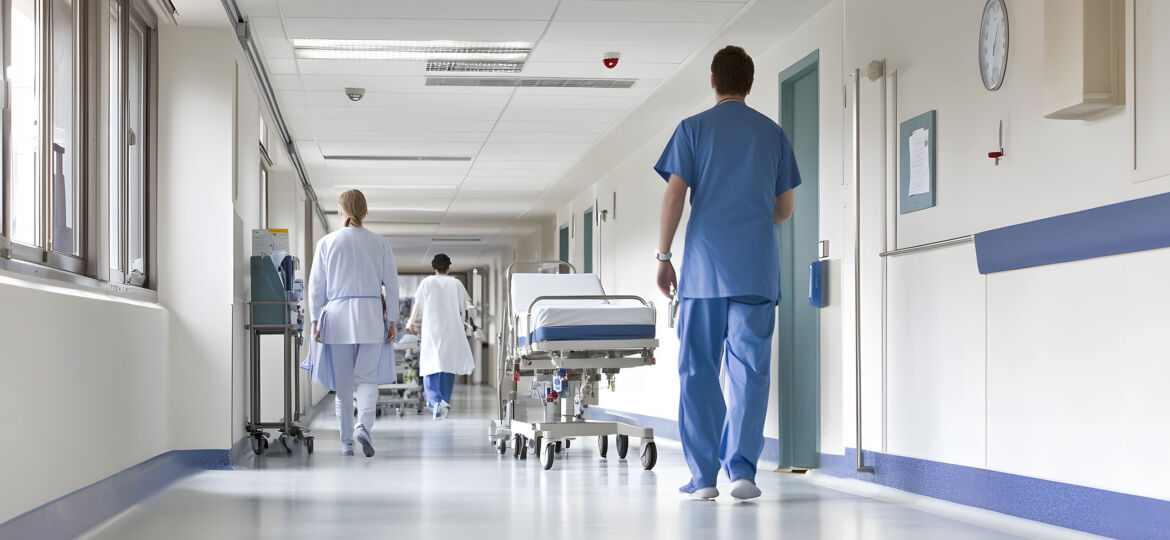 A bright hospital corridor with medical staff walking in various directions. Two healthcare professionals in blue scrubs and one in a white coat are visible, along with a patient bed being pushed by a staff member. The hallway features clean white walls, large windows on the left, and a wall clock. Medical equipment and charts are visible on the walls.