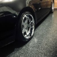 Close-up of a black sports car's shiny chrome wheel and side panel, parked on a textured grey garage floor. The car's body reflects the overhead lighting, highlighting its polished surface and sleek design.