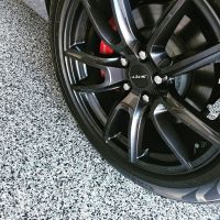Close-up of a black sports car's alloy wheel with a distinctive 'ZXS' logo, showcasing its multi-spoke design and red brake caliper, against a textured grey and white speckled floor.