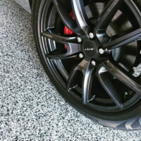 Close-up of a black sports car's alloy wheel with a distinctive 'ZXS' logo, showcasing its multi-spoke design and red brake caliper, against a textured grey and white speckled floor.