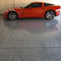 Bright red sports car parked on a glossy, polished concrete floor with a speckled finish, reflecting the vehicle's silhouette. The garage door is partially visible in the background.