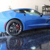 A vibrant blue sports car with sleek bodywork and black rims with red detailing is parked on a glossy epoxy garage floor with speckles. The reflective surface of the floor creates a mirror-like effect, highlighting the car's well-maintained condition. Visible in the background are garage storage units and equipment, suggesting a clean and organized space.