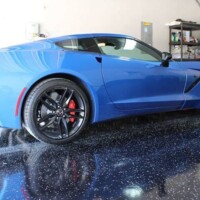 A vibrant blue sports car with sleek bodywork and black rims with red detailing is parked on a glossy epoxy garage floor with speckles. The reflective surface of the floor creates a mirror-like effect, highlighting the car's well-maintained condition. Visible in the background are garage storage units and equipment, suggesting a clean and organized space.