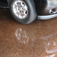 A close-up of a speckled brown epoxy garage floor partially wet from rain, showing the reflection of a silver car's tire and bumper. The wet spots on the floor create a glossy effect contrasting with the dry, textured appearance of the epoxy coating.