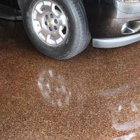 A close-up of a speckled brown epoxy garage floor partially wet from rain, showing the reflection of a silver car's tire and bumper. The wet spots on the floor create a glossy effect contrasting with the dry, textured appearance of the epoxy coating.