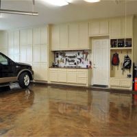 A well-organized garage with a glossy stained concrete floor, featuring a black pickup truck parked on the left. The right side shows a wall with built-in cream cabinets, a workbench with various tools hanging above it, a step ladder, and a couple of backpacks hanging on hooks. A green ball and a bucket are on the floor to the right, indicating a space used for both storage and activities.
