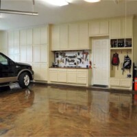 A well-organized garage with a glossy stained concrete floor, featuring a black pickup truck parked on the left. The right side shows a wall with built-in cream cabinets, a workbench with various tools hanging above it, a step ladder, and a couple of backpacks hanging on hooks. A green ball and a bucket are on the floor to the right, indicating a space used for both storage and activities.