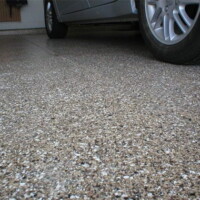 Close-up view of a textured concrete driveway with scattered pebbles, leading to a car's silver front wheel and part of the vehicle visible at the top of the image.
