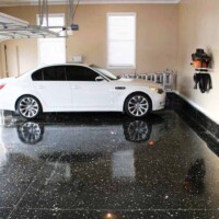 View from the entrance of an empty garage with a grey speckled epoxy floor. The garage door opener is mounted on the ceiling with the door partially raised. A white side door with a security keypad is visible on the right, and the walls are painted a light neutral color.