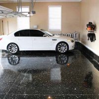 View from the entrance of an empty garage with a grey speckled epoxy floor. The garage door opener is mounted on the ceiling with the door partially raised. A white side door with a security keypad is visible on the right, and the walls are painted a light neutral color.