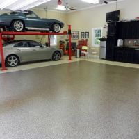 Spacious garage with a multi-tone speckled floor. A silver sports car is parked underneath a classic car on a red car lift. The garage includes a black cabinet set, a small dining area, and decorative items such as a vintage Coca-Cola cooler