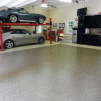 Spacious garage with a multi-tone speckled floor. A silver sports car is parked underneath a classic car on a red car lift. The garage includes a black cabinet set, a small dining area, and decorative items such as a vintage Coca-Cola cooler
