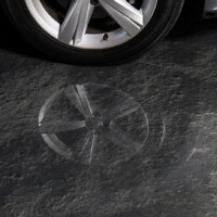 Close-up of a dark textured garage floor showing a clear, reflective watermark shaped like the rim of a car wheel, with the actual car wheel and tire partially visible in the upper left corner.