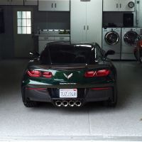 A dark green Corvette parked inside a well-organized garage with speckled flooring. The garage is equipped with white storage cabinets, a washer and dryer set, and a red motorcycle parked to the right. Natural light filters through a window on the garage door.