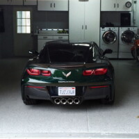 A dark green Corvette parked inside a well-organized garage with speckled flooring. The garage is equipped with white storage cabinets, a washer and dryer set, and a red motorcycle parked to the right. Natural light filters through a window on the garage door.
