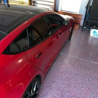 A bright red sedan parked on a speckled pink and gray epoxy-coated garage floor. The car features a sleek design with tinted windows, and black rims. The garage is organized, with a wooden workbench and tool cabinet to the left, and a closed white door at the far end, suggesting a clean and well-maintained personal automotive space.