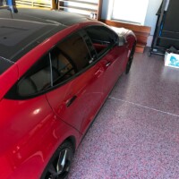 A bright red sedan parked on a speckled pink and gray epoxy-coated garage floor. The car features a sleek design with tinted windows, and black rims. The garage is organized, with a wooden workbench and tool cabinet to the left, and a closed white door at the far end, suggesting a clean and well-maintained personal automotive space.