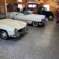 Inside a well-lit, wooden garage are three classic cars parked on a smooth, polished concrete floor. Closest to the camera is a cream convertible with a closed top, followed by a white convertible with a red interior and open top, and at the back, a black vintage truck with yellow-rimmed wheels. The garage features exposed wood beams, string lights, an American flag, and a tidy tool storage area, creating a nostalgic and cozy atmosphere for automobile enthusiasts.